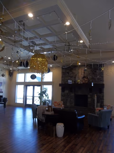 A spacious living room area with wooden flooring, a stone fireplace, and large windows letting in natural light. The ceiling is decorated with hanging gold and black ornaments and string lights. There are several chairs and a small table arranged near the fireplace.