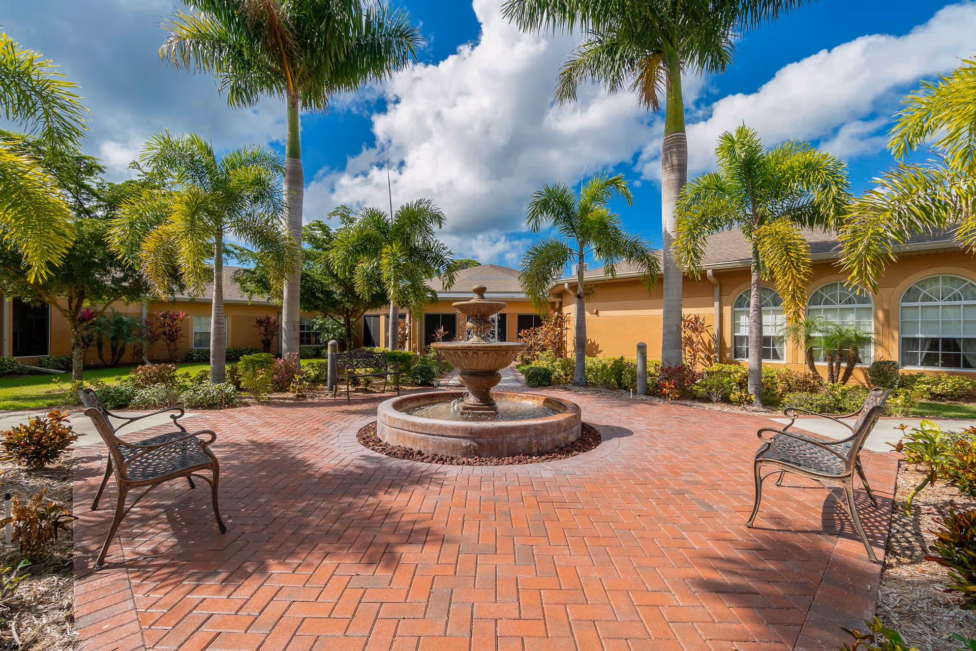 Sunlit courtyard with a central tiered fountain, metal benches, palm trees, and a single-story building in the background.