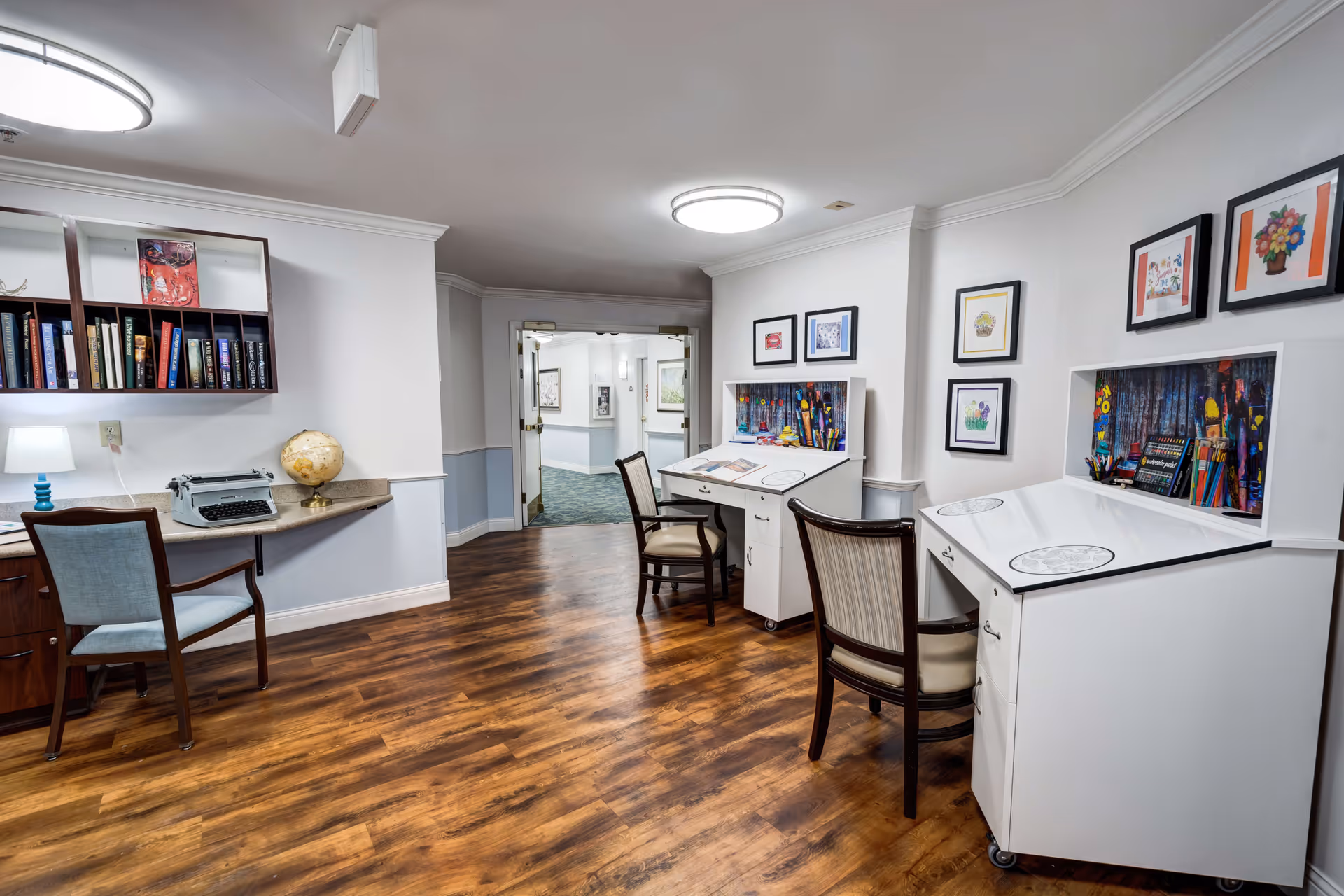 Interior room with wooden flooring featuring two white art desks with chairs, colorful art supplies, and framed artwork on the walls. On the left side, there is a wooden desk with a chair, a globe, a typewriter, and a bookshelf with books and decorative items. The room is well-lit with ceiling lights and has a doorway leading to a hallway.