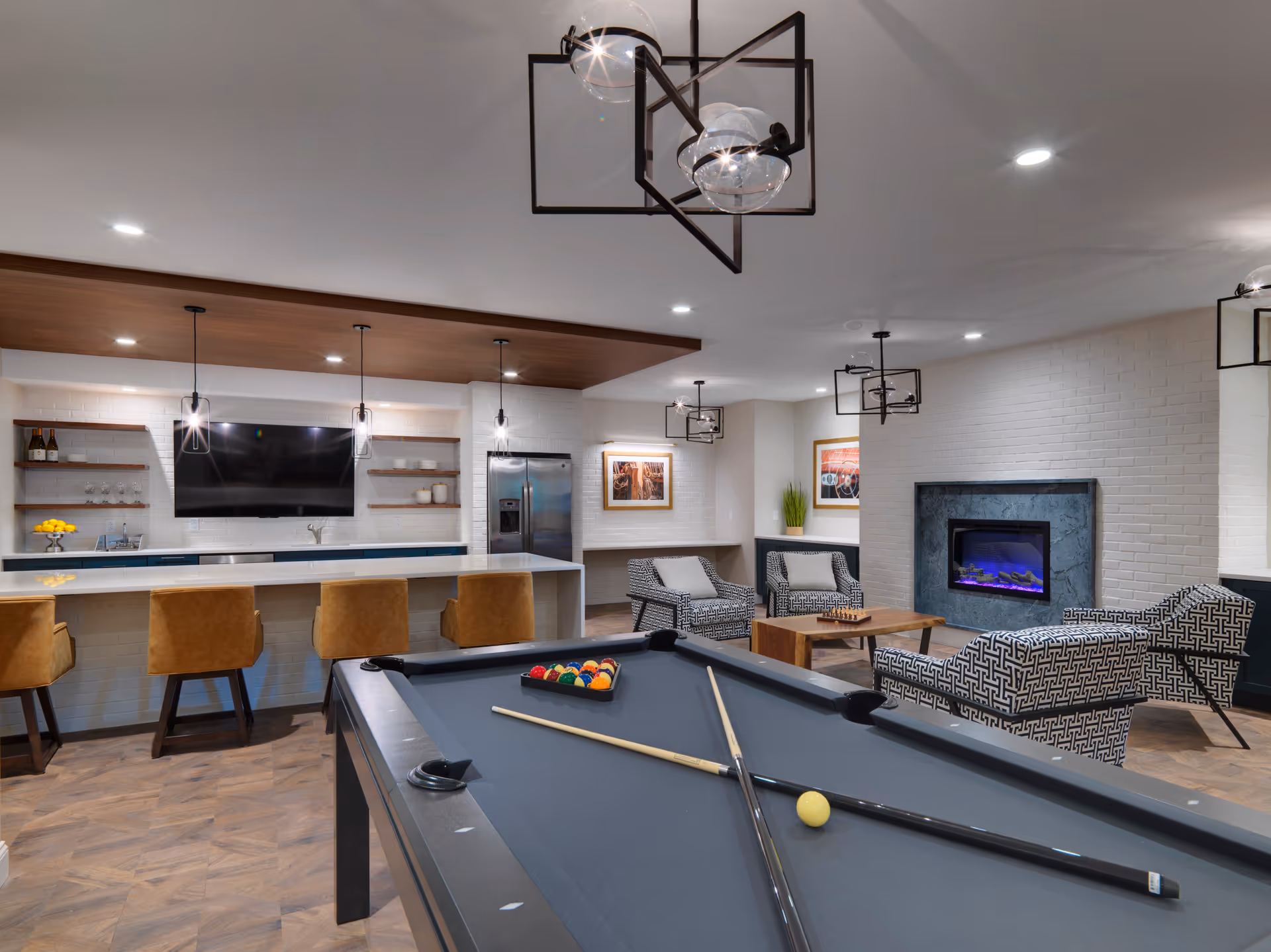 A modern recreational room featuring a pool table with cues and balls, a seating area with patterned armchairs around a wooden coffee table, and a fireplace with a blue flame. In the background, there is a kitchen area with a long counter, four brown bar stools, a wall-mounted TV, open shelves with glasses and bottles, and a stainless steel refrigerator. The room has contemporary pendant lighting and wood-patterned flooring.