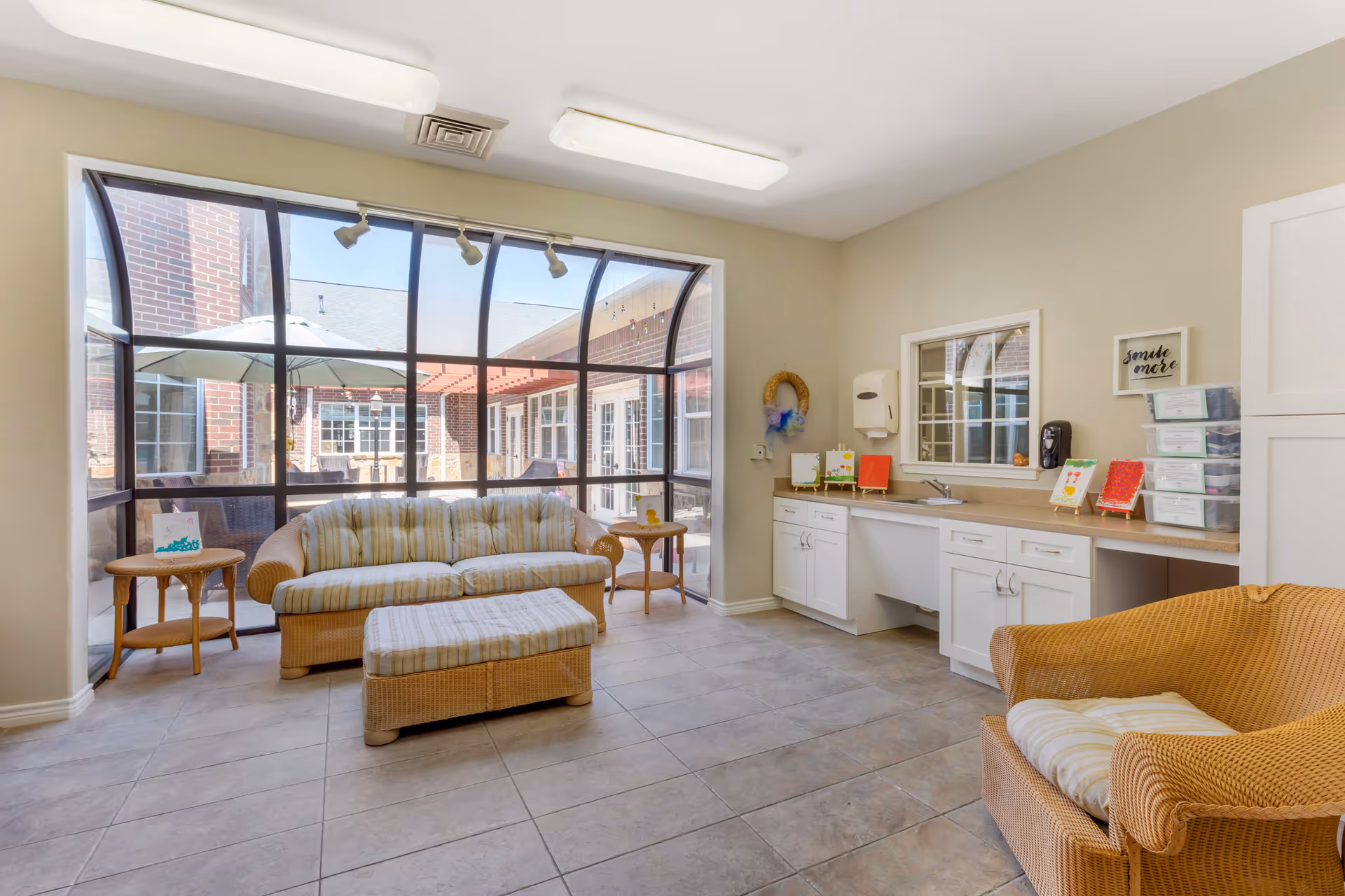 A bright and clean sitting area with wicker furniture including a striped cushioned sofa, matching ottoman, and armchair. The room has large curved windows providing a view of an outdoor patio with an umbrella and seating. There are white cabinets with a countertop, a sink, and small decorative items on the right side. The floor is tiled and the walls are painted light beige.