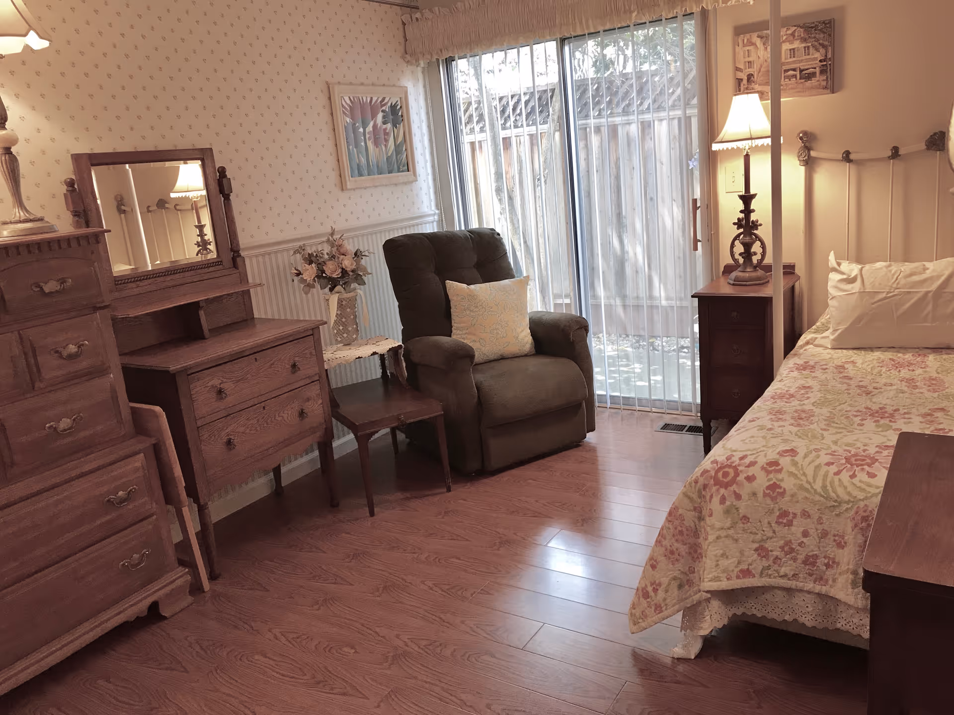 A cozy bedroom with wooden furniture including a dresser with a mirror, a nightstand with a lamp, and a bed with floral bedding. There is a green upholstered armchair with a decorative pillow next to a small table holding a vase of flowers. A sliding glass door with vertical blinds leads to an outdoor area.
