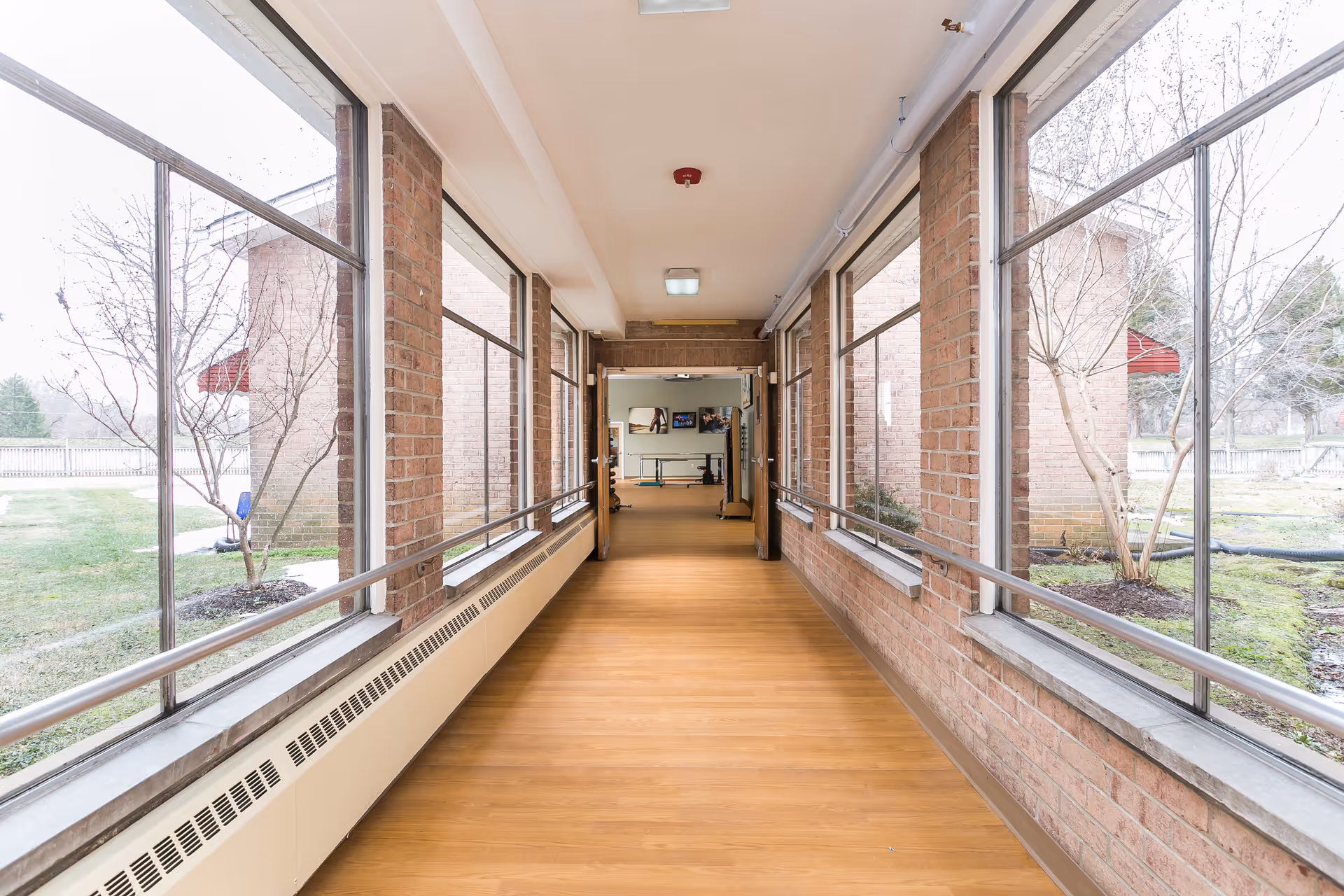 Bright indoor corridor with wood-look flooring, handrails and large windows overlooking a courtyard in a nursing facility.