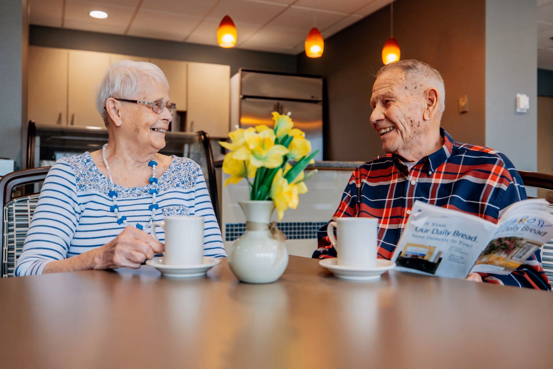 An elderly woman and man sitting at a table in a kitchen or dining area, smiling and enjoying a conversation. The woman is wearing glasses and a striped shirt with a blue necklace, holding a white mug. The man is wearing a red and blue plaid shirt, holding a newspaper and a white mug. A vase with yellow flowers is on the table between them, and pendant lights hang from the ceiling above.