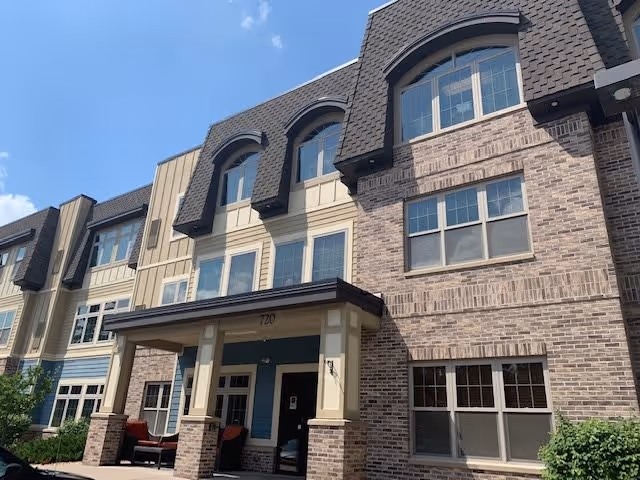Exterior view of a multi-story assisted living facility building with a mix of brick and siding facade, large windows, and a covered entrance with seating. The sky is clear and blue.