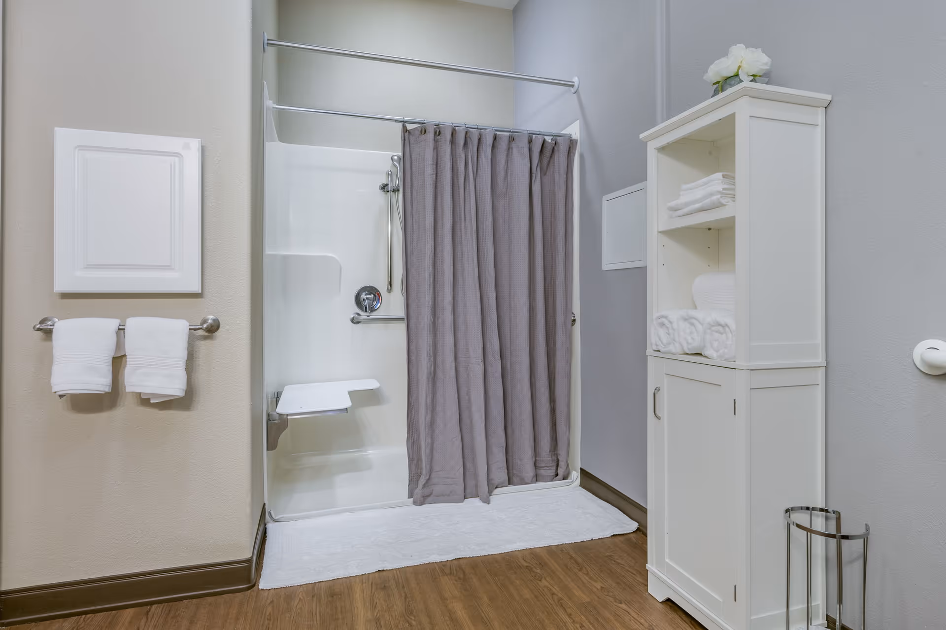 A clean and accessible bathroom shower area with a white built-in shower seat, a gray shower curtain, and a white bath mat on a wooden floor. On the left wall, there is a towel rack with two white towels, and on the right side, a white cabinet with shelves holding neatly folded white towels and a decorative flower on top.