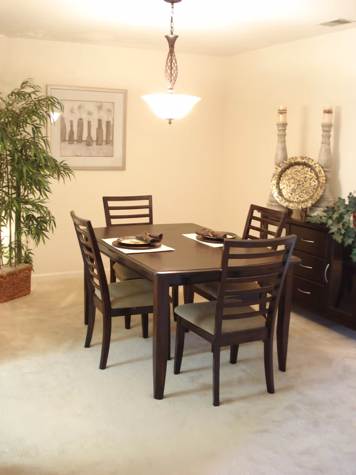 A dining room with a dark wooden rectangular table and four matching chairs with cushioned seats. The table is set with placemats, plates, and napkins. There is a hanging light fixture above the table, a framed artwork on the wall, a potted plant in a wicker basket, and a dark wooden sideboard with decorative items including two tall candle holders and a decorative plate.