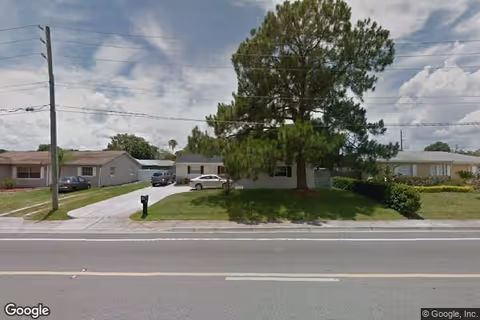 Single-story house with a large pine tree in the front yard, a driveway with parked cars, and a mailbox by the road.