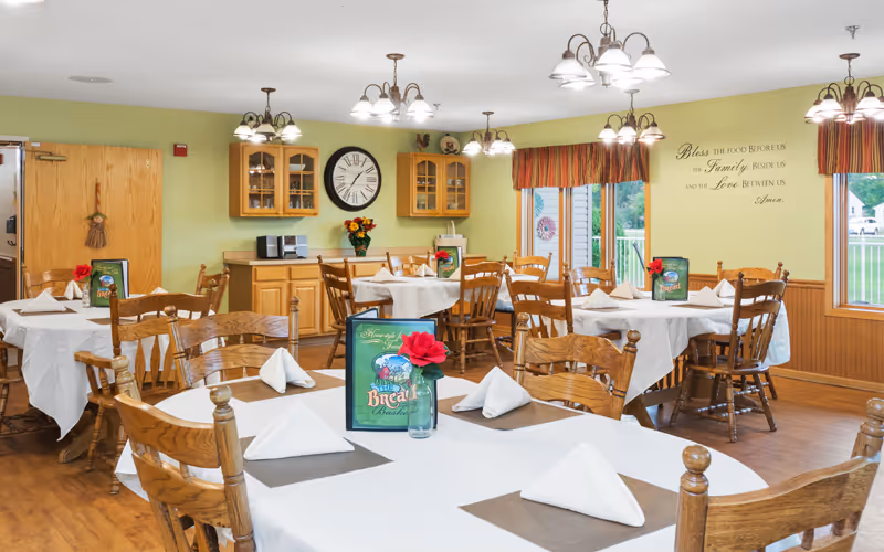 A dining room with several wooden tables and chairs arranged neatly. Each table is covered with a white tablecloth, set with folded white napkins, placemats, and a small vase holding a red flower. The walls are painted light green with wooden wainscoting, and there are windows with striped valances letting in natural light. A large clock and wooden cabinets are mounted on the wall, and a decorative wall decal reads, 'Bless the food before us, the family beside us, and the love between us. Amen.'