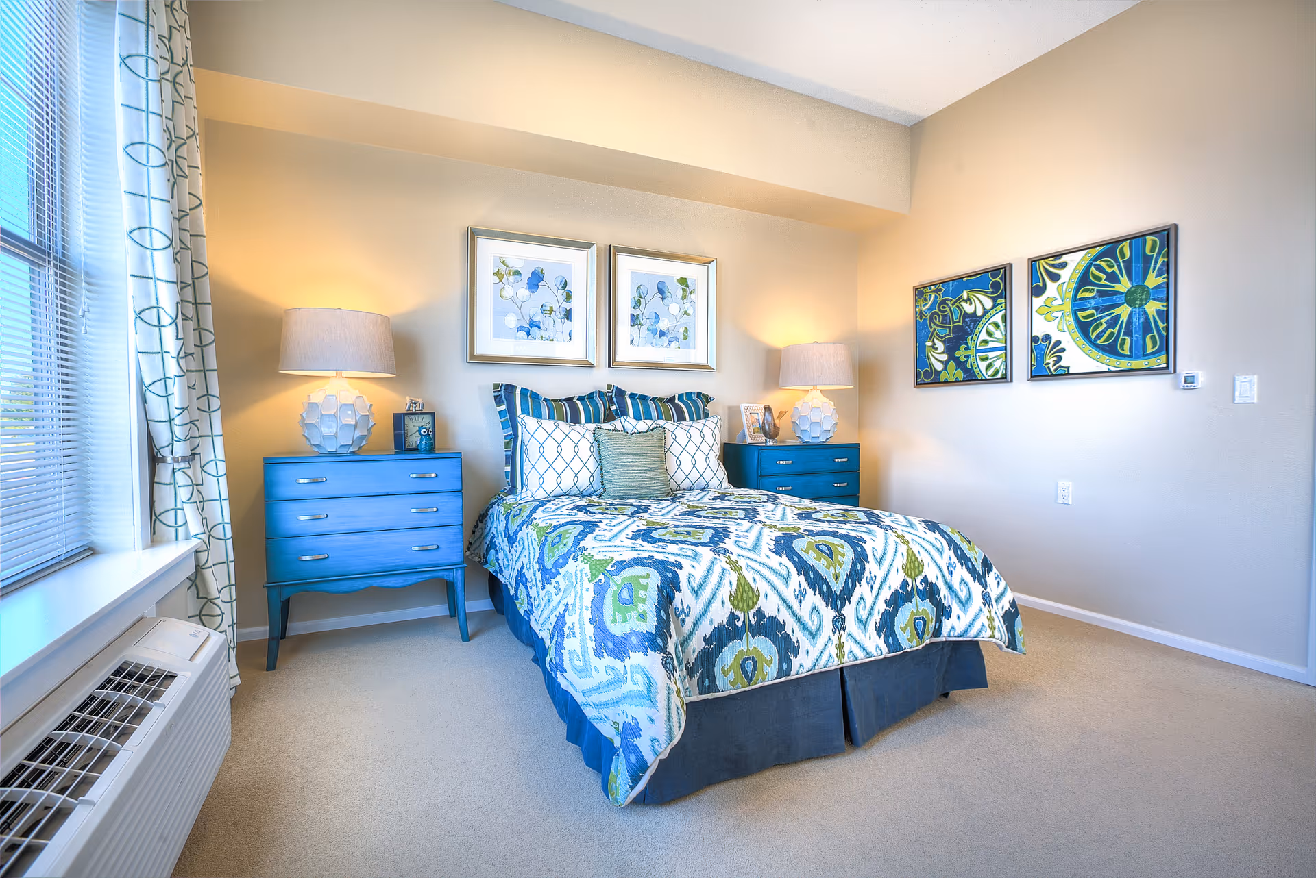 A bright and cozy bedroom with a bed featuring a blue, green, and white patterned comforter and multiple pillows. There are two blue nightstands on either side of the bed, each with a white lamp. Two framed floral artworks hang above the bed, and two additional colorful framed artworks are on the adjacent wall. A window with patterned curtains is on the left side, and a white air conditioning unit is below the window.