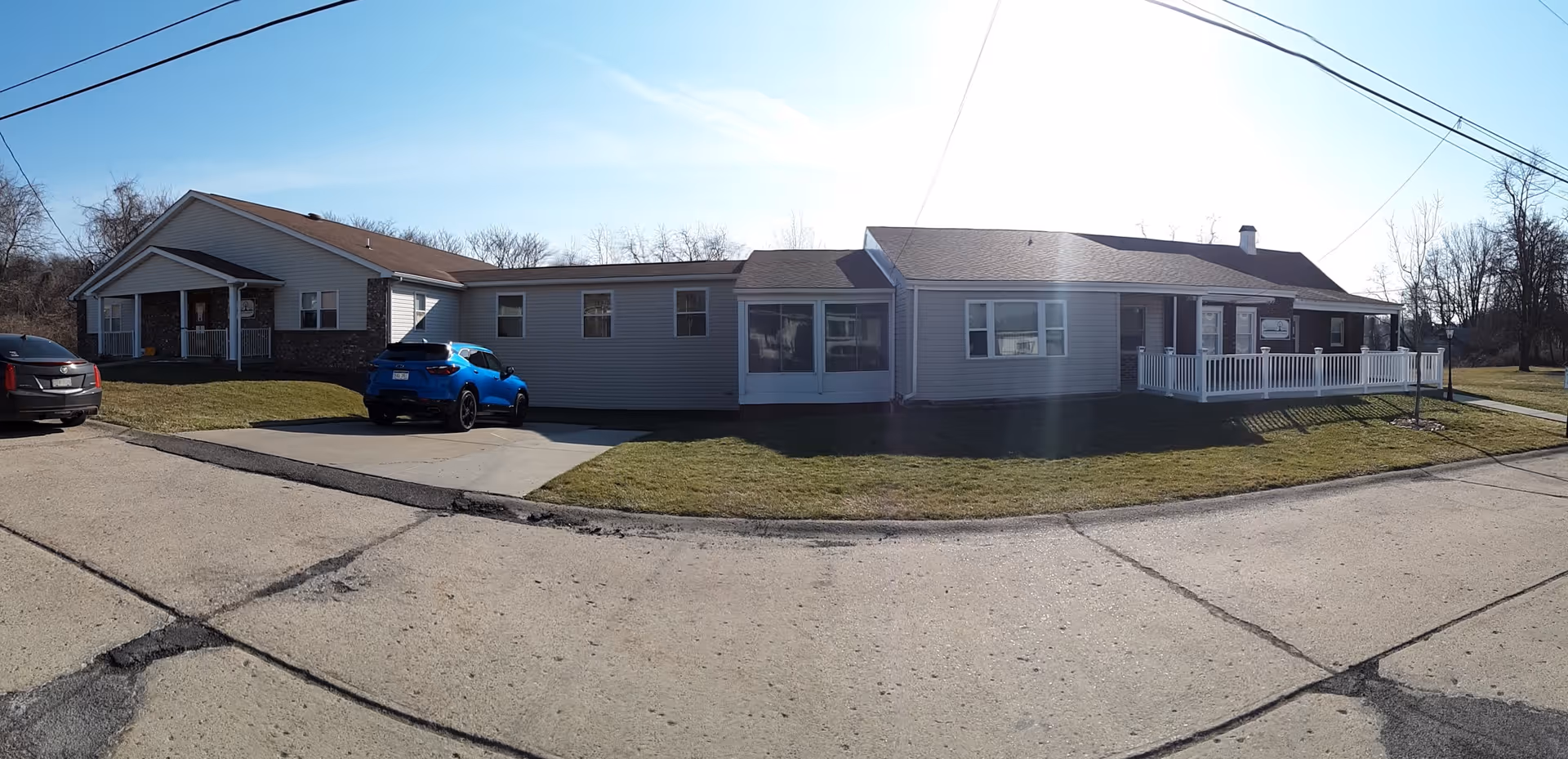 Exterior view of a single-story assisted living facility building with beige siding and a brown roof. There are two cars parked in front on a paved driveway. The building has a small porch with white railings and a sign near the entrance. The surrounding area includes a grassy lawn and leafless trees in the background under a clear sky.