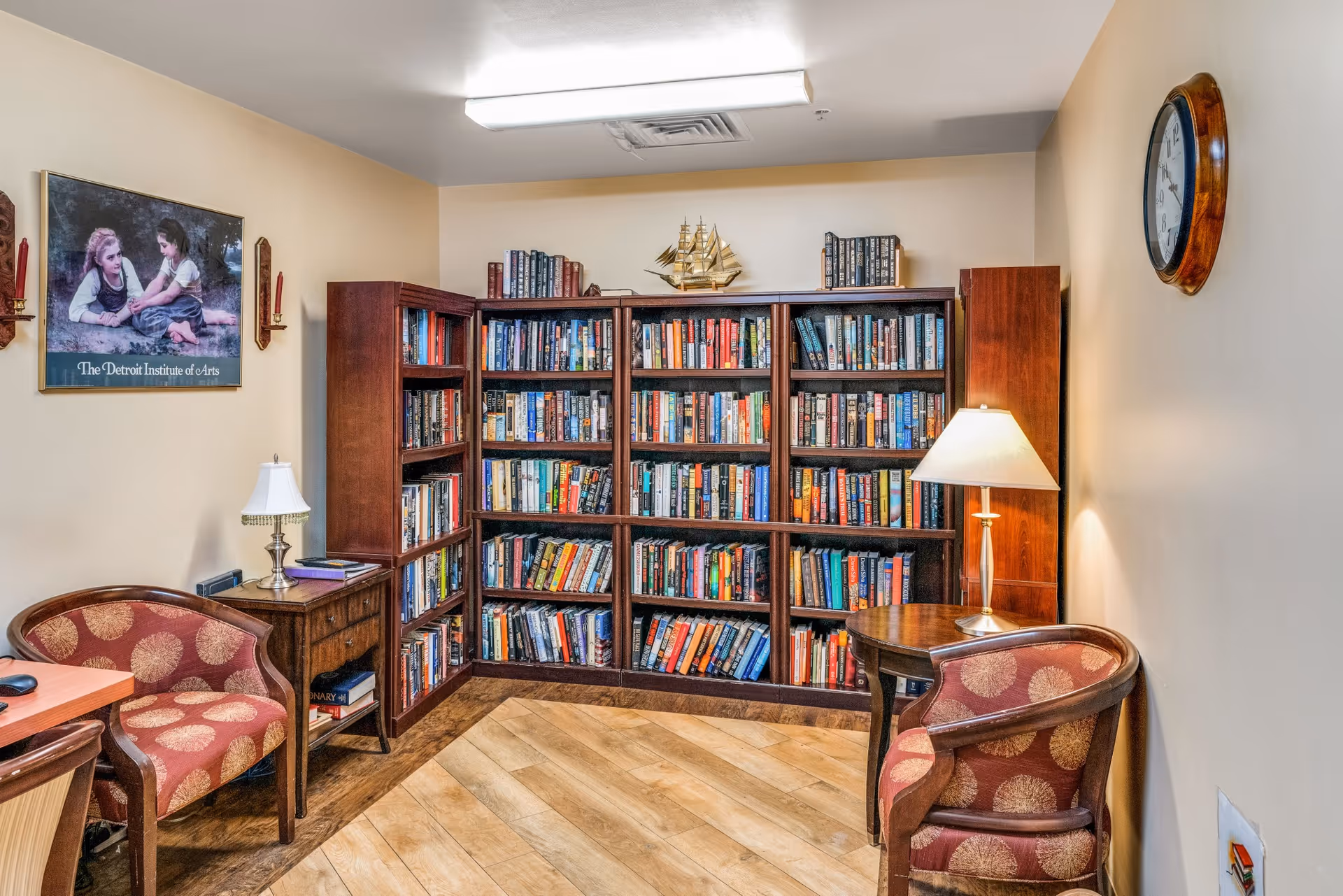 Cozy interior reading room with wooden bookshelves filled with books, two upholstered chairs, side tables with lamps, and wall art.
