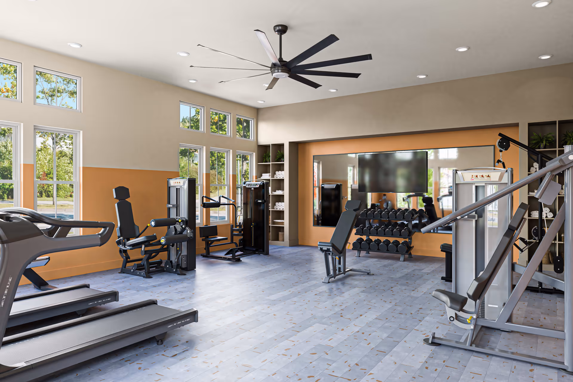 A bright and spacious fitness room with large windows letting in natural light. The room contains various exercise equipment including treadmills, weight machines, a bench, and a rack of dumbbells. There is a large ceiling fan and a wall-mounted TV above the dumbbell rack. Shelves with towels and plants are visible in the background.