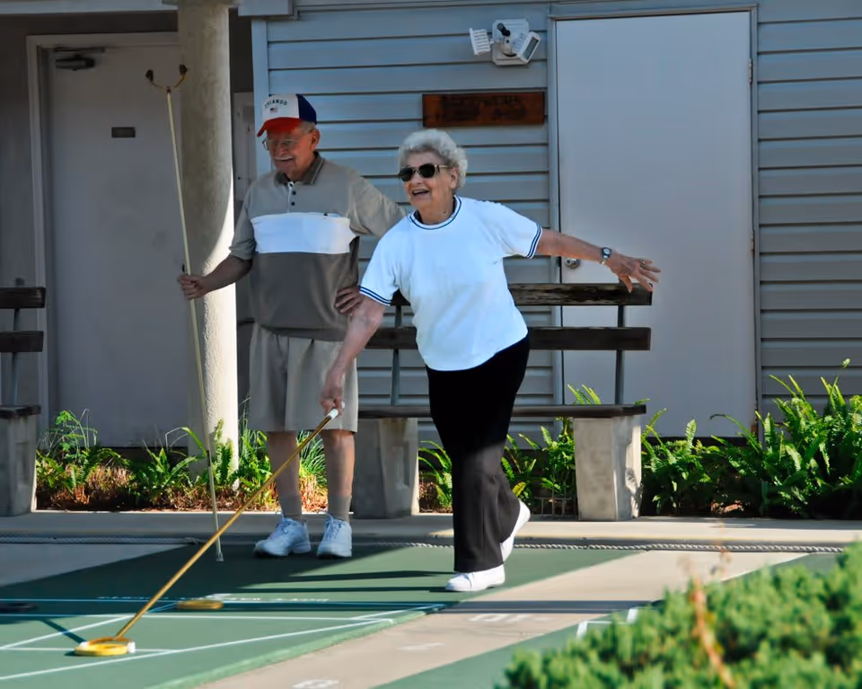 An elderly woman wearing sunglasses and a white shirt is playing shuffleboard outdoors, while an elderly man stands behind her holding a shuffleboard cue. They are on a shuffleboard court with benches and plants in the background.