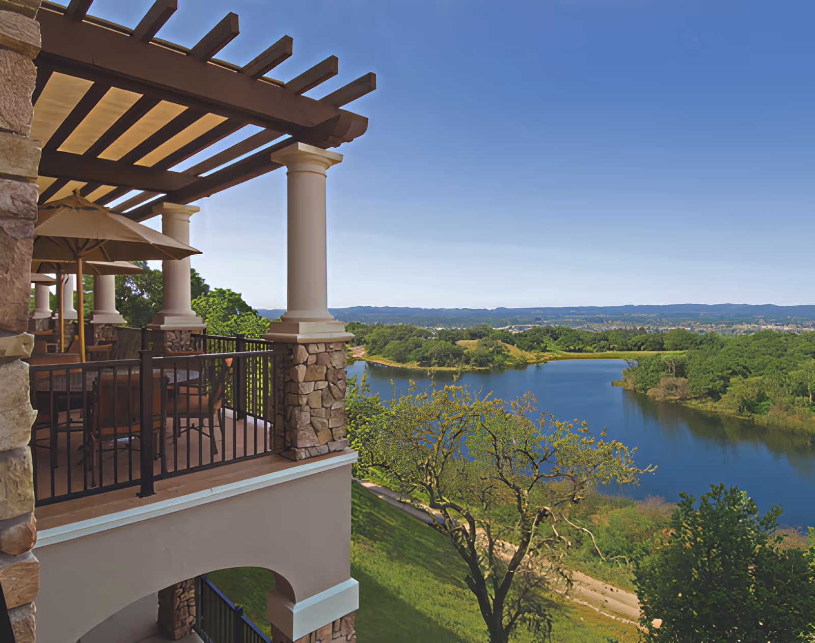 A covered balcony with stone columns and patio seating overlooking a lake and surrounding green landscape under a clear blue sky.