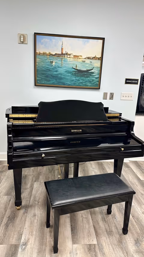 A black Kohler piano with a matching black bench in front of it, placed on a wood-patterned floor. Above the piano, there is a framed painting depicting a waterfront scene with boats and buildings under a partly cloudy sky. A small sign on the wall to the right reads 'DINING ROOM'.