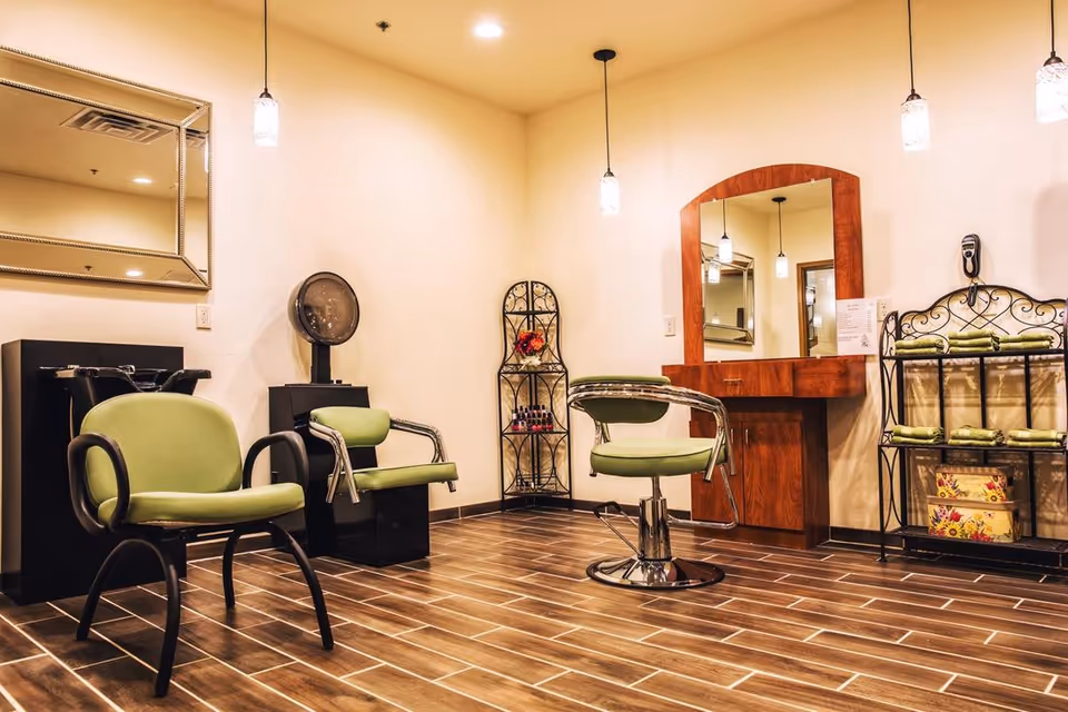 A bright salon-style room with green styling chairs, mirrors, pendant lights, and shelving on a wood-look tile floor.