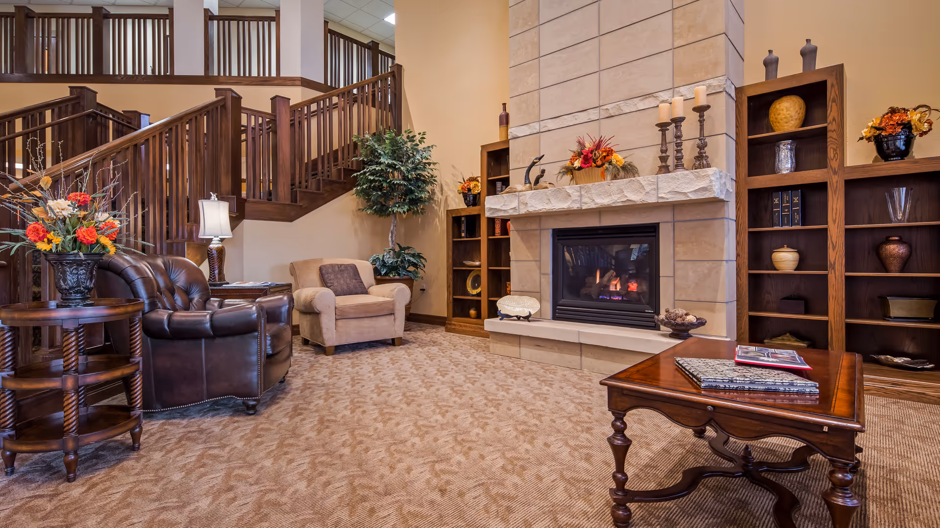 Cozy living room area with a stone fireplace, wooden bookshelves decorated with vases and candles, a brown leather armchair, a beige armchair, a wooden coffee table with books, and a wooden side table with a floral arrangement. A staircase with wooden railings is visible in the background.