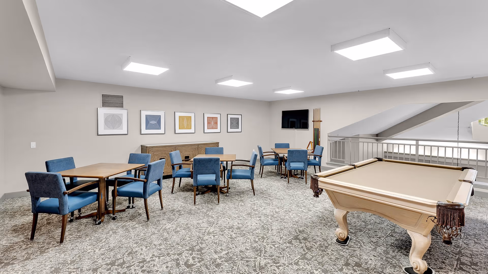 A spacious common area with several tables and blue upholstered chairs arranged for group seating. A beige pool table is positioned on the right side of the room. The walls are decorated with framed abstract art, and a flat-screen TV is mounted on the far wall. The room has bright overhead lighting and a patterned carpeted floor.