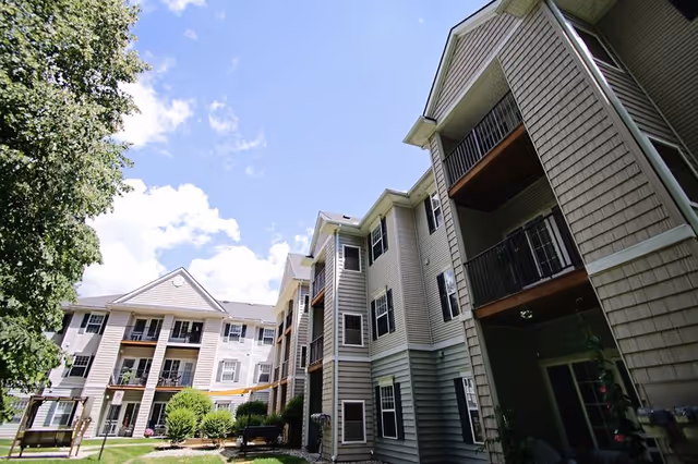 Exterior view of a multi-story senior living facility building with balconies, surrounded by greenery and trees under a partly cloudy blue sky.