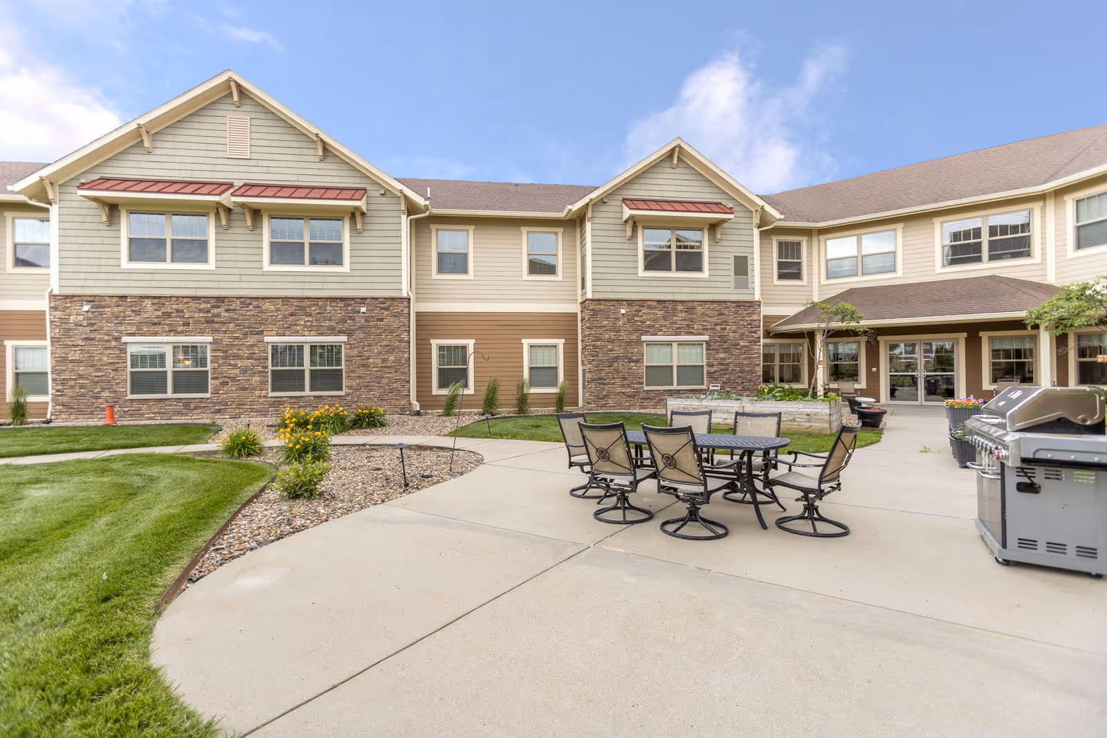 Outdoor patio area at Independence Village of Ankeny featuring a round table with six chairs, a barbecue grill, and a two-story building with stone and siding exterior in the background under a partly cloudy sky.