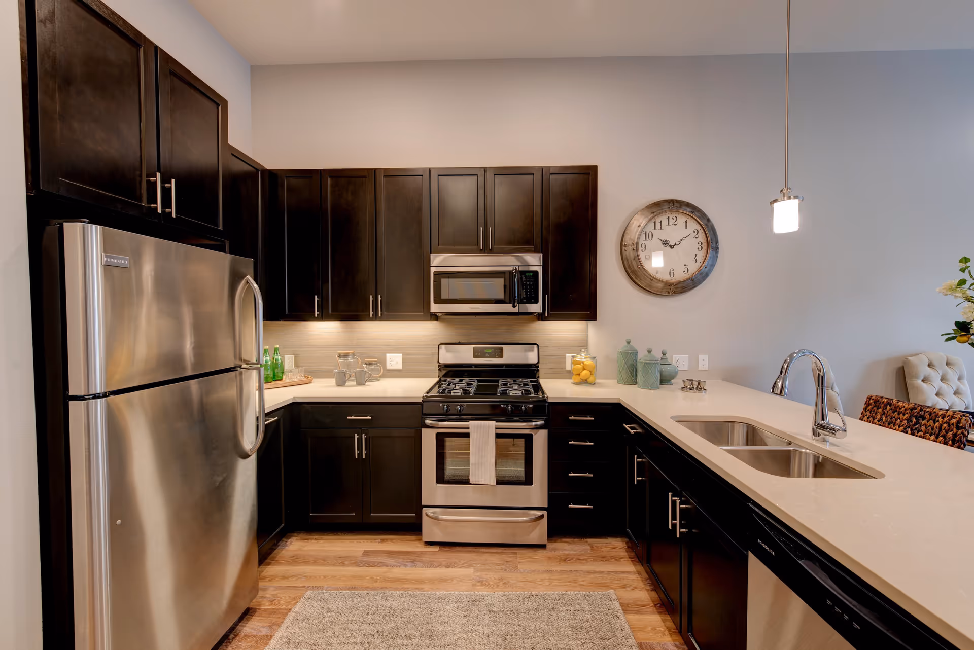 Modern kitchen with dark wood cabinets, stainless steel refrigerator, stove, microwave, and dishwasher. The kitchen features a light-colored countertop with a double sink, a wall clock, and decorative jars with lemons and other items. There is a pendant light hanging above the counter and a beige rug on the wooden floor.