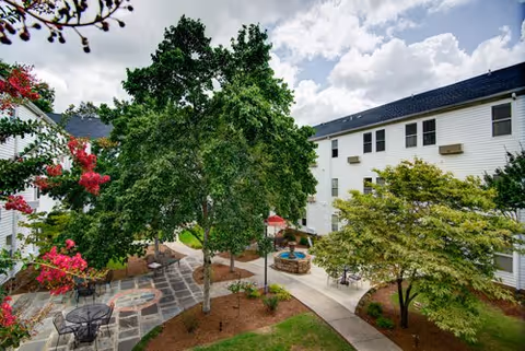 Outdoor courtyard area at Morningside of Vestavia Hills featuring large trees, a paved walkway, patio tables with chairs, a small fountain, and surrounding white residential buildings under a partly cloudy sky.