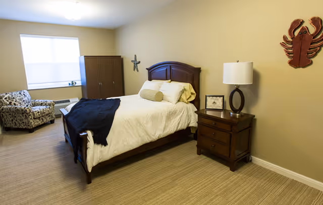 A cozy bedroom with a wooden bed frame, white bedding, and a dark throw blanket. There is a wooden nightstand with a lamp and a framed picture on it. A patterned armchair is placed near a window with a white blind. A wooden wardrobe stands against the wall, and decorative wall hangings including a starfish and a lobster are visible.