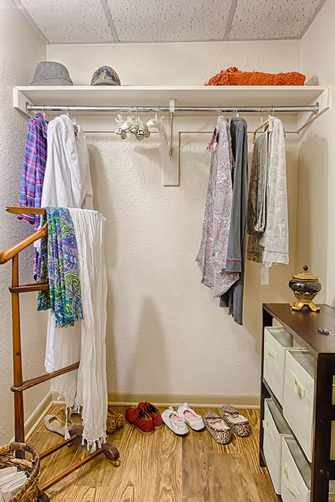 A small closet area with a hanging rod holding various clothing items including shirts and robes. On the top shelf, there are hats and a folded orange blanket. Below, several pairs of shoes are neatly arranged on a wooden floor. To the left, a wooden rack holds scarves and a white cloth. On the right, a dark wooden storage unit with fabric drawers is visible, topped with a decorative item.