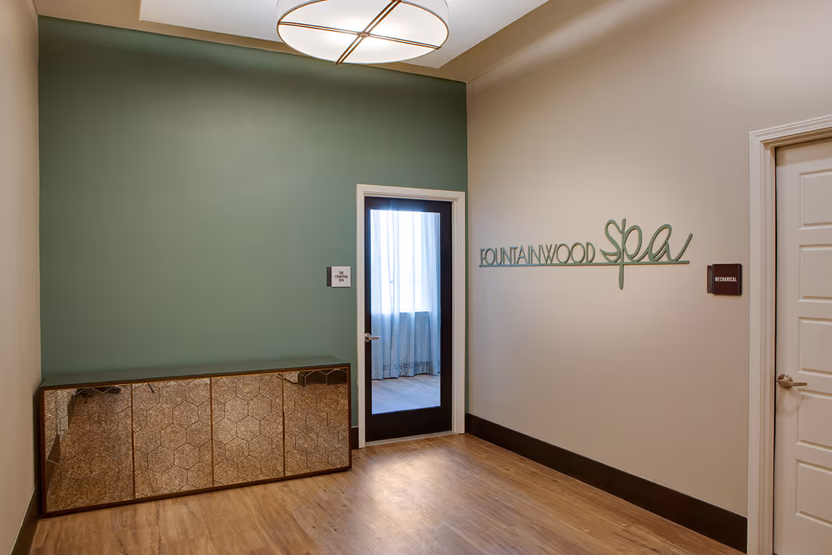 Interior hallway with wooden flooring and a green accent wall. A door with a glass panel and curtain leads to the Fountainwood Spa. There is a decorative cabinet with a reflective surface against the green wall. On the opposite wall, the words 'FOUNTAINWOOD spa' are mounted in green lettering. Another door labeled 'MECHANICAL' is visible on the right side.