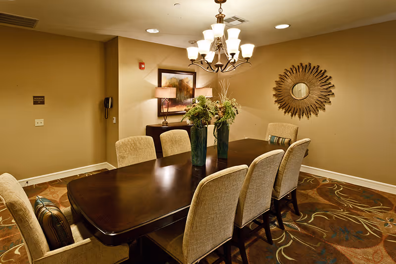 A dining room with a dark wooden rectangular table surrounded by eight beige upholstered chairs. Two green vases with floral arrangements are placed on the table. The room features a patterned carpet, a chandelier with multiple lights hanging from the ceiling, a framed painting, a table lamp on a sideboard, and a decorative sunburst mirror on the wall.
