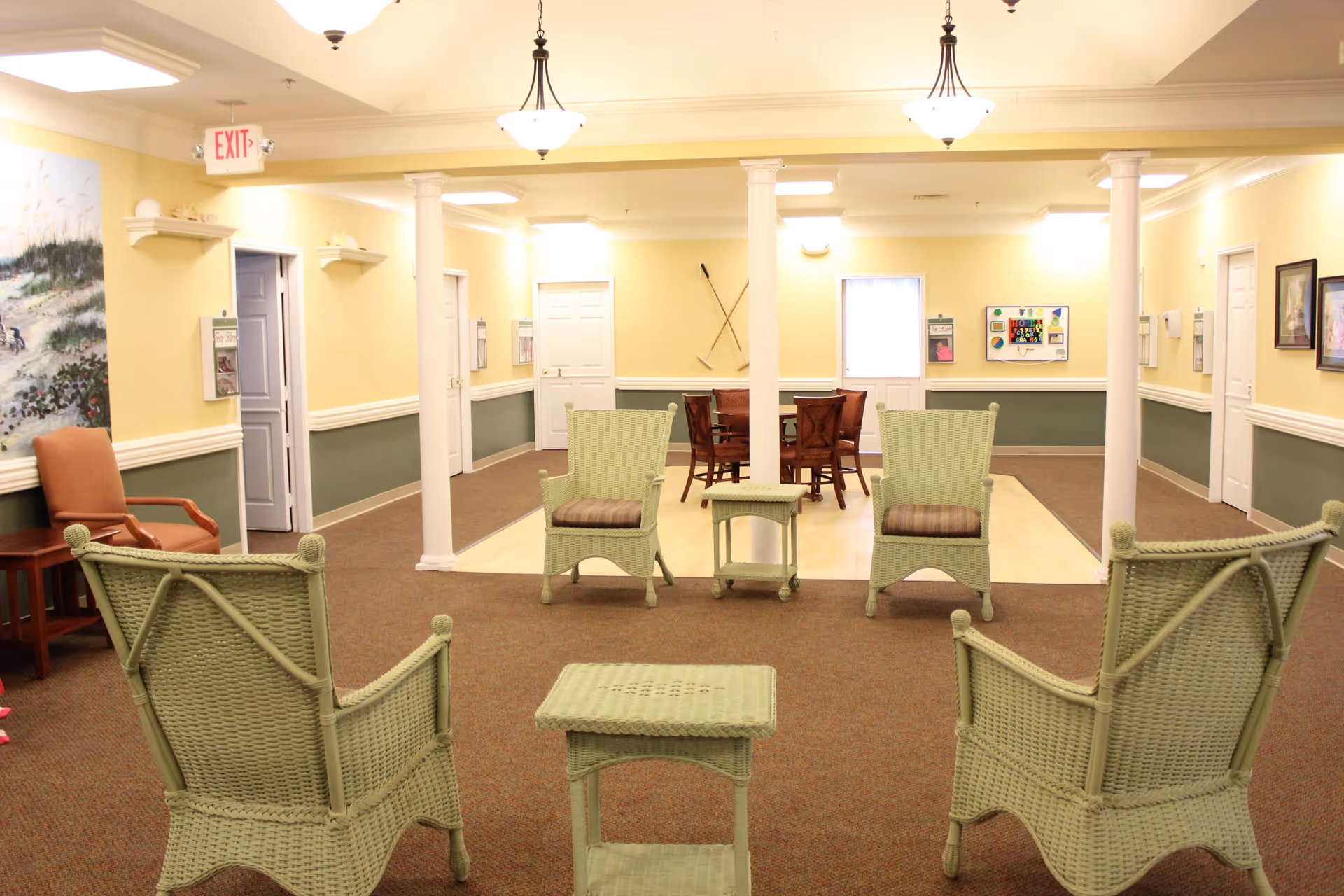 A well-lit common area in a senior living facility with light yellow walls and green wainscoting. The room features four green wicker chairs arranged around two small matching tables in the foreground, and a wooden table with four chairs in the background. The space has white columns, ceiling lights, framed artwork, and an exit sign above a door on the left side.