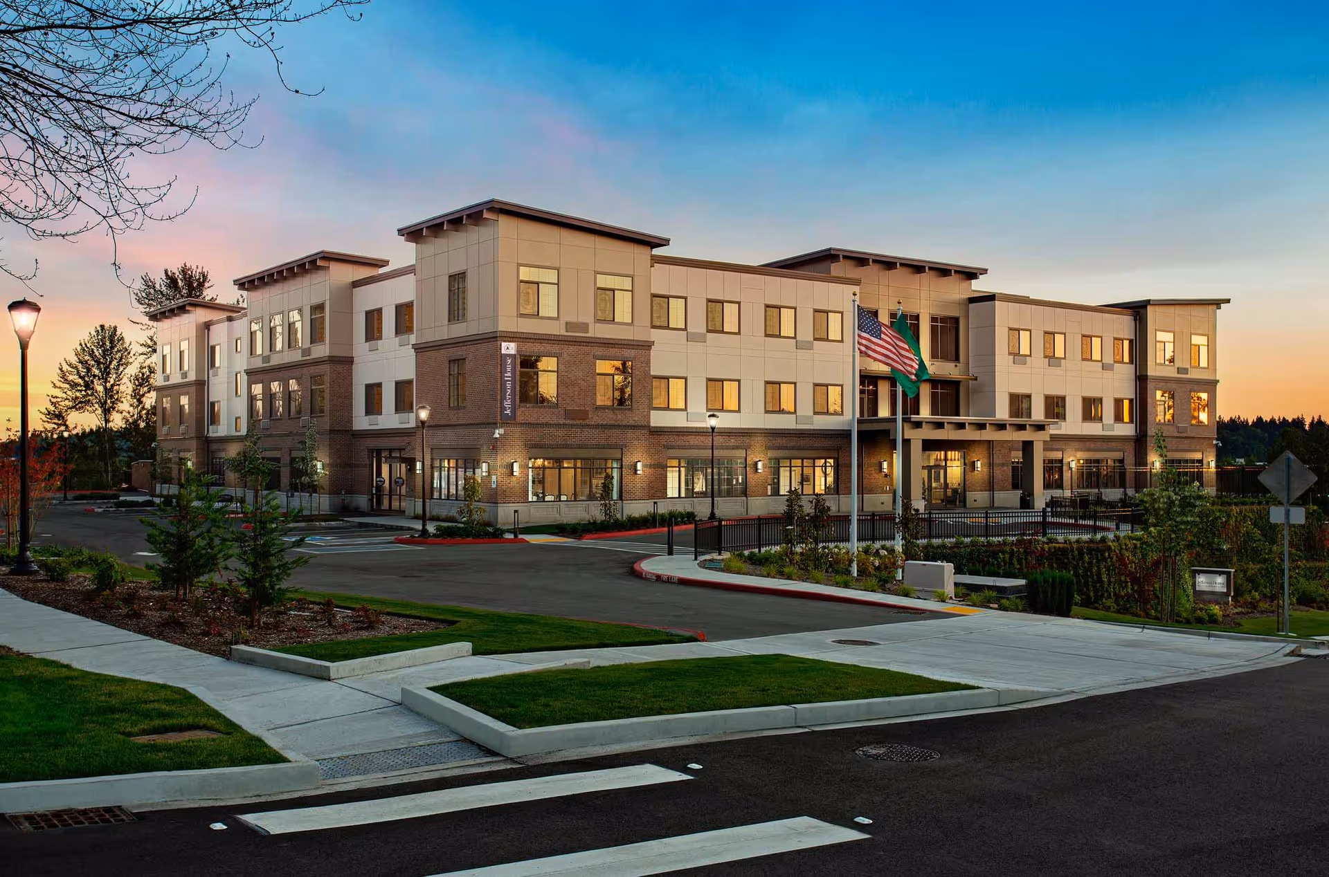 Exterior view of a modern three-story building at sunset with well-maintained landscaping, street lamps, and two flagpoles displaying the American flag and another flag in front of the entrance.