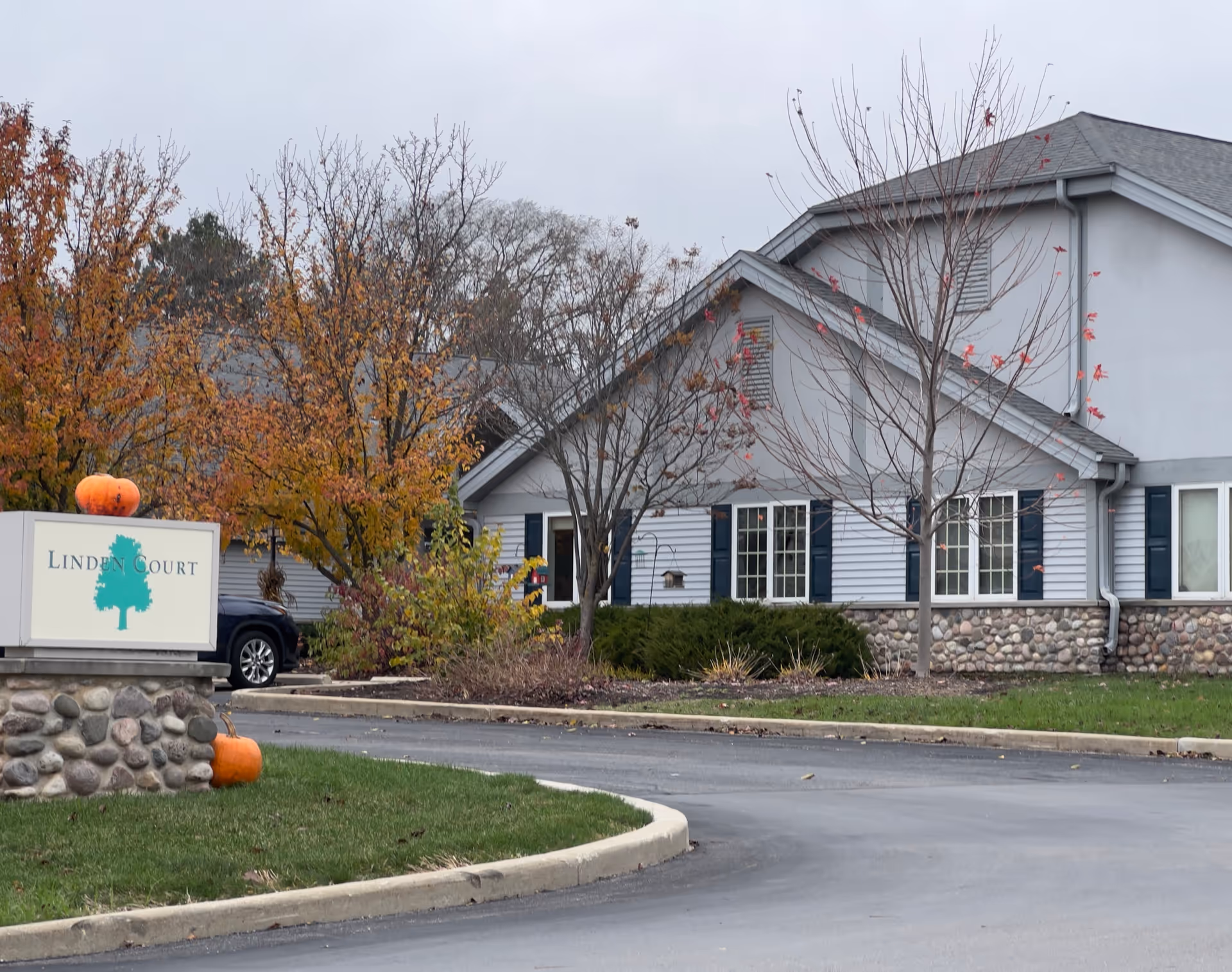 Exterior view of Linden Court Waukesha facility with a stone and siding building, autumn trees with colorful leaves, a curved driveway, and a sign displaying the facility name with pumpkins placed on and near the sign.