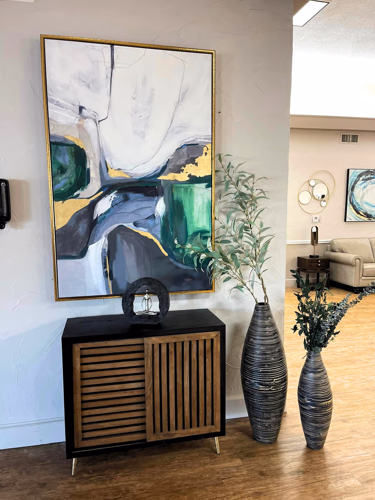 A corner of an interior space featuring a wooden cabinet with slatted doors, a modern abstract painting with green, white, gray, and gold tones hanging above it, and two tall decorative vases with green foliage placed on the wooden floor. In the background, a beige sofa, a side table with a lamp, and circular wall mirrors are visible.