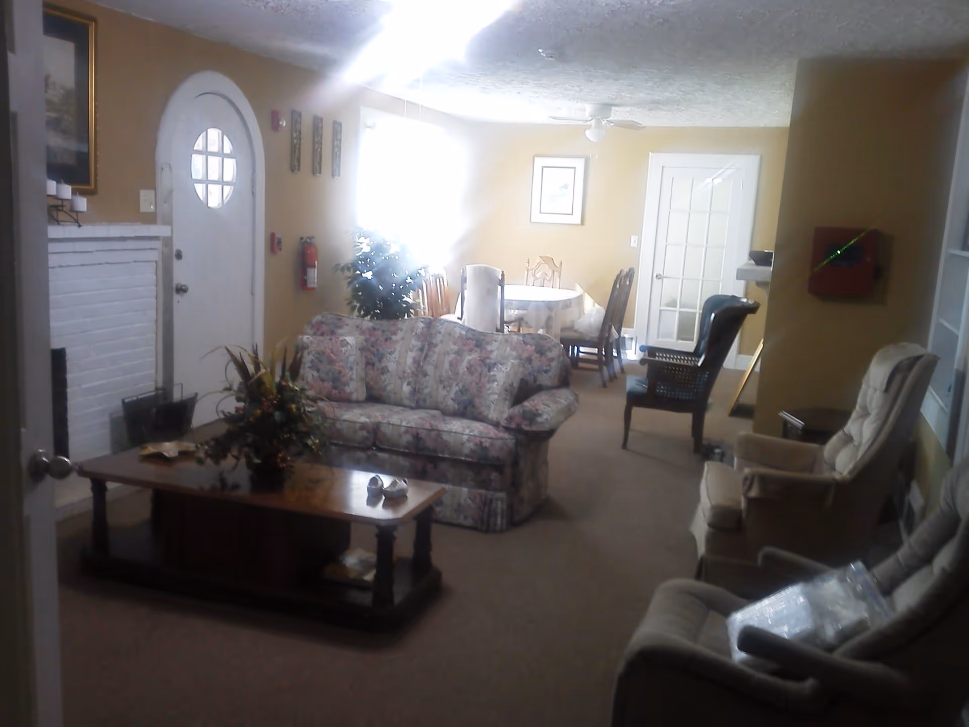A cozy living room area with a floral patterned sofa, a wooden coffee table with a flower arrangement, and several armchairs. In the background, there is a dining table with chairs near a window letting in bright light. The walls are painted beige, and there is a white door with a circular window. A white brick fireplace is visible on the left side.