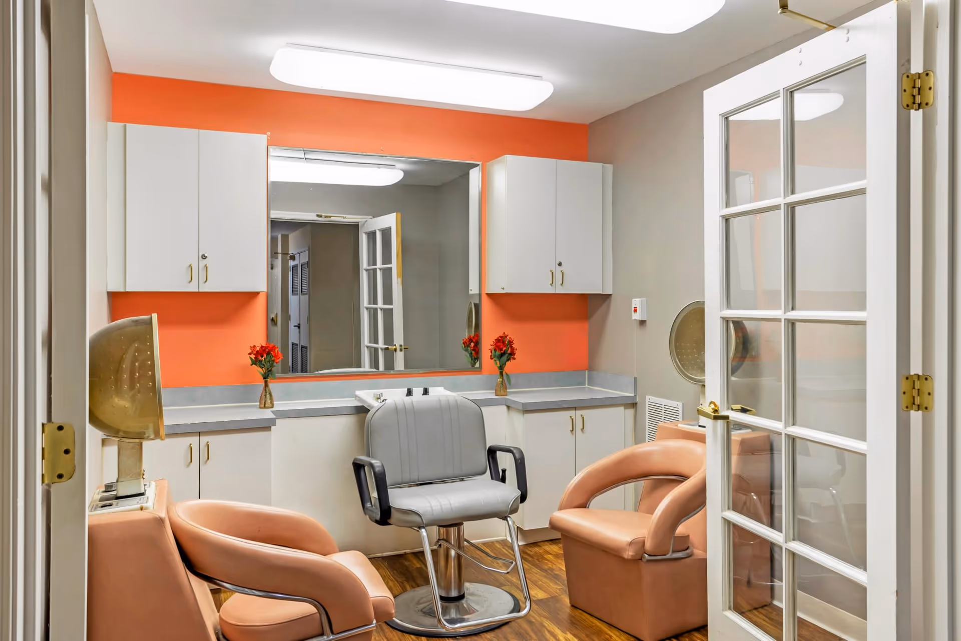 Interior view of a small salon or grooming room with two peach-colored salon chairs and one gray salon chair in front of a large mirror. The walls are painted gray with an orange accent behind the mirror. There are white cabinets above and below the countertop, and two small vases with red flowers are placed on the counter. A hair dryer hood is visible on the left side. The room has wooden flooring and a glass-paneled door on the right.