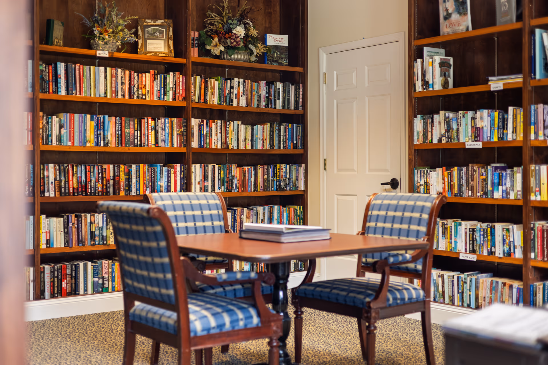 A cozy library room with wooden bookshelves filled with books lining the walls. In the center, there is a wooden table with a book on it, surrounded by four cushioned chairs upholstered in blue and beige plaid fabric. The room has a beige carpet and a white door in the background.