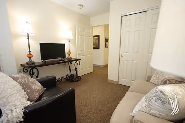 A cozy living room area with a beige sofa and a black armchair, both adorned with decorative pillows. A wooden console table with ornate metal legs holds a flat-screen TV and two table lamps with white shades. The room has beige walls and carpeted flooring, with a partially open door leading to a hallway and a closed closet with sliding doors.