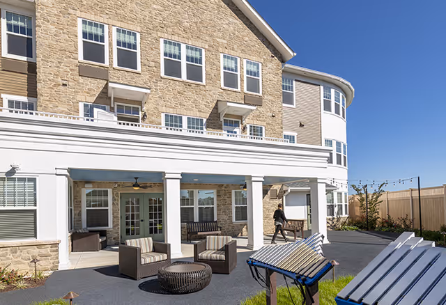 Outdoor patio area of a senior living facility with comfortable seating including cushioned chairs and a round table under a covered porch supported by white columns. A person is walking near the porch, and there are musical chimes and a large xylophone in the foreground. The building has a stone facade with multiple windows and a clear blue sky above.