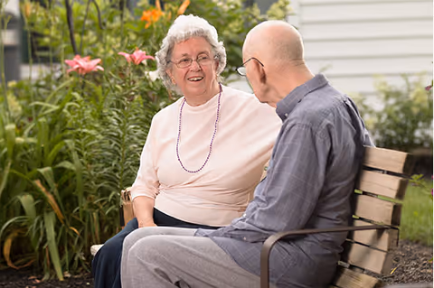 An elderly woman and an elderly man sitting on a wooden bench outdoors, engaged in conversation. The woman is wearing glasses, a light pink long-sleeve top, and a beaded necklace. The man is wearing glasses and a gray checkered shirt. Behind them are green plants and flowers, with a white building in the background.