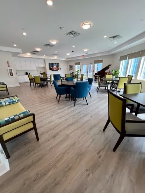 A spacious, well-lit common area with multiple tables and chairs arranged for seating. The room features light wood flooring, a piano near large windows with blinds, and a wall-mounted television. There are also some plants and a built-in counter with cabinets along one wall.