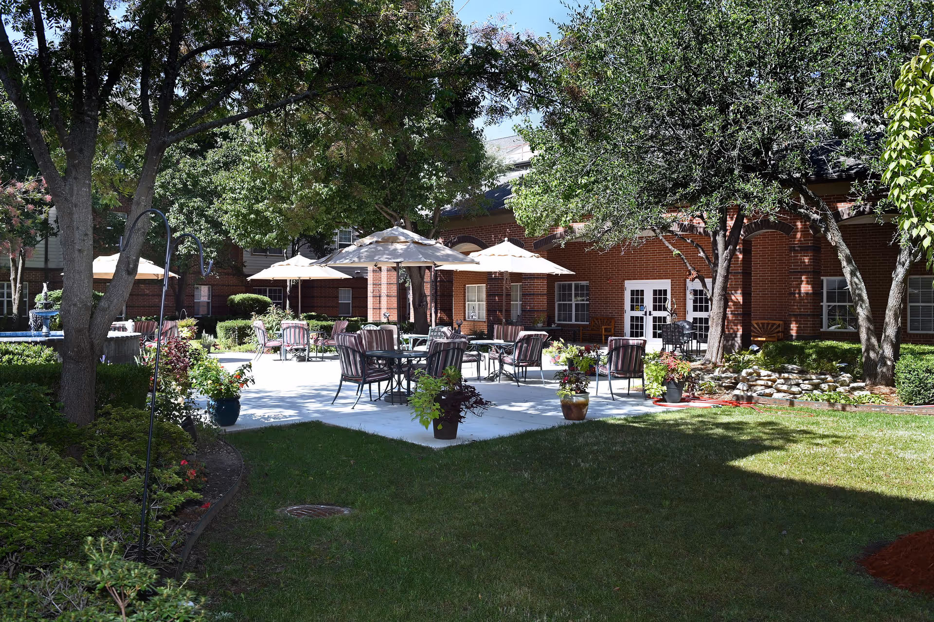 Outdoor patio area at Town Village Tulsa featuring multiple tables with striped cushioned chairs and beige umbrellas. The patio is surrounded by green grass, trees, potted plants, and a brick building in the background with white doors and windows.