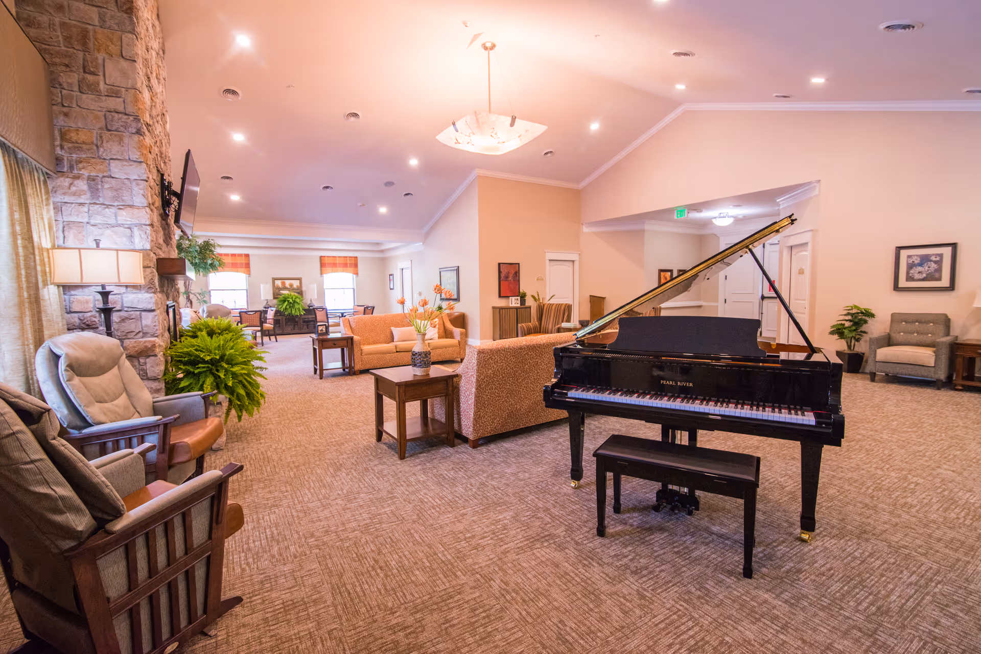 A spacious and well-lit living room area in a senior living facility featuring a black grand piano with a matching bench in the center. The room has beige carpeting, comfortable seating including armchairs and sofas, a stone fireplace with a mounted TV, several potted plants, and framed artwork on the walls. Large windows with curtains allow natural light to brighten the space.