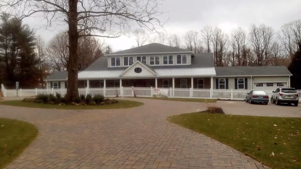 Front exterior of a single-story care facility with a white picket fence, covered porch, brick driveway, and parked cars.