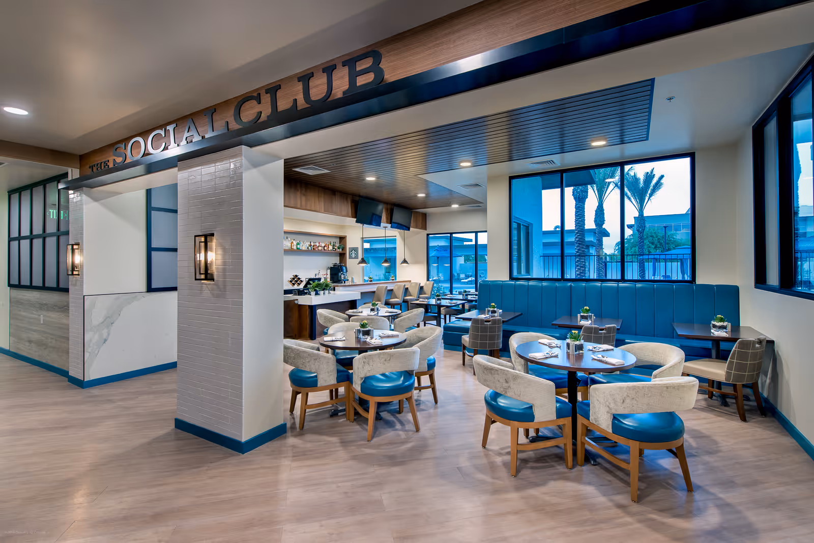Interior view of a modern social club dining area with round tables, cushioned chairs with blue seats, and a long blue banquette along the window. The space features large windows showing palm trees outside, a bar area with stools, and warm lighting under a wooden ceiling panel. The entrance has a sign above reading 'THE SOCIAL CLUB'.