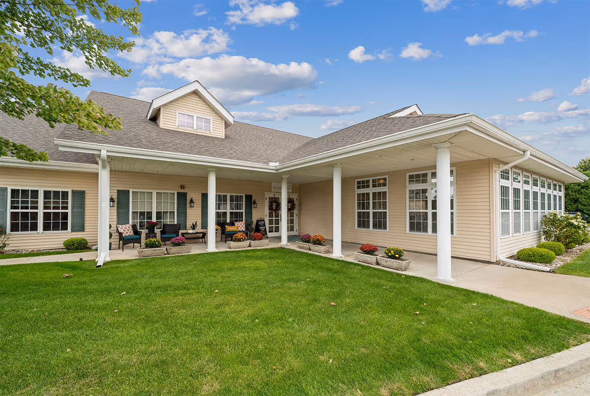 Exterior view of a single-story beige building with white columns supporting a covered porch area. There are several windows with white frames and green shutters, outdoor seating with chairs and small tables, and flower pots with colorful flowers along the porch. The building is surrounded by a well-maintained green lawn and a partly cloudy blue sky is visible above.