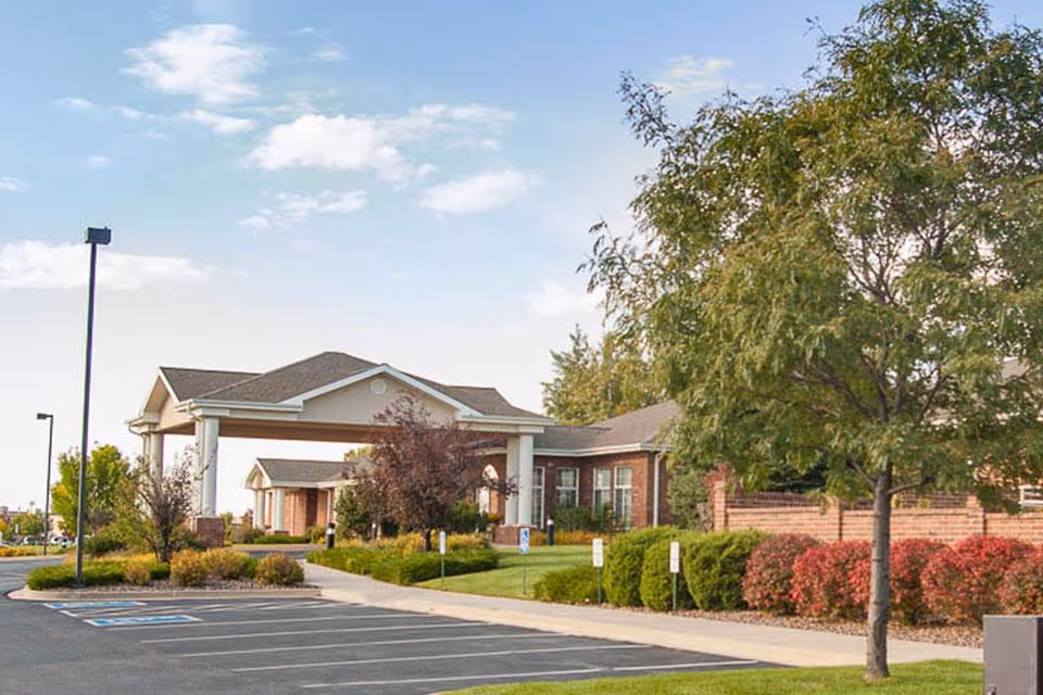 Front exterior of a single-story brick senior living facility with a covered entrance, landscaped grounds, and a parking lot.