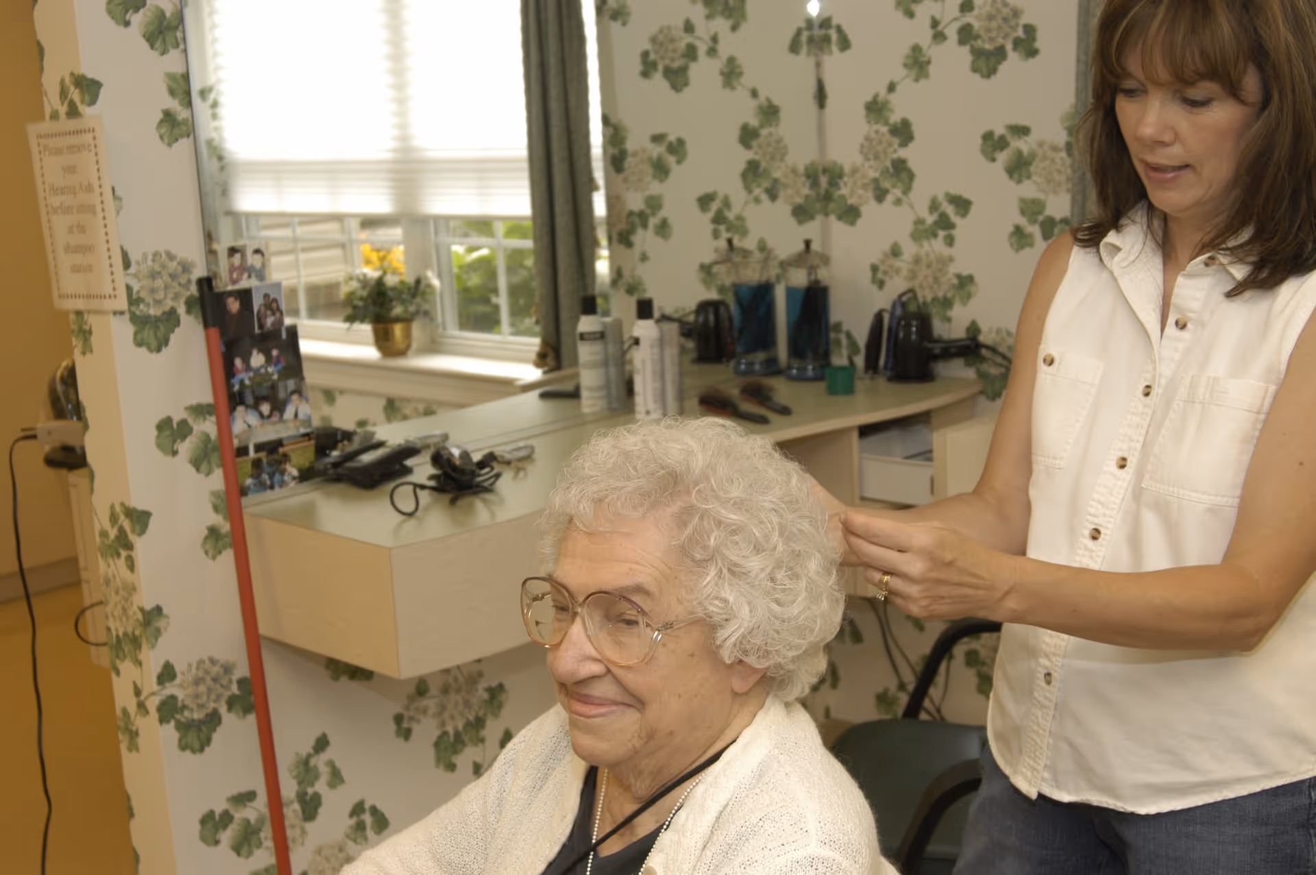 An elderly woman with curly white hair and glasses is seated while a younger woman styles her hair in a room with floral wallpaper and a window with blinds. The room has a counter with hair care products and a mirror.