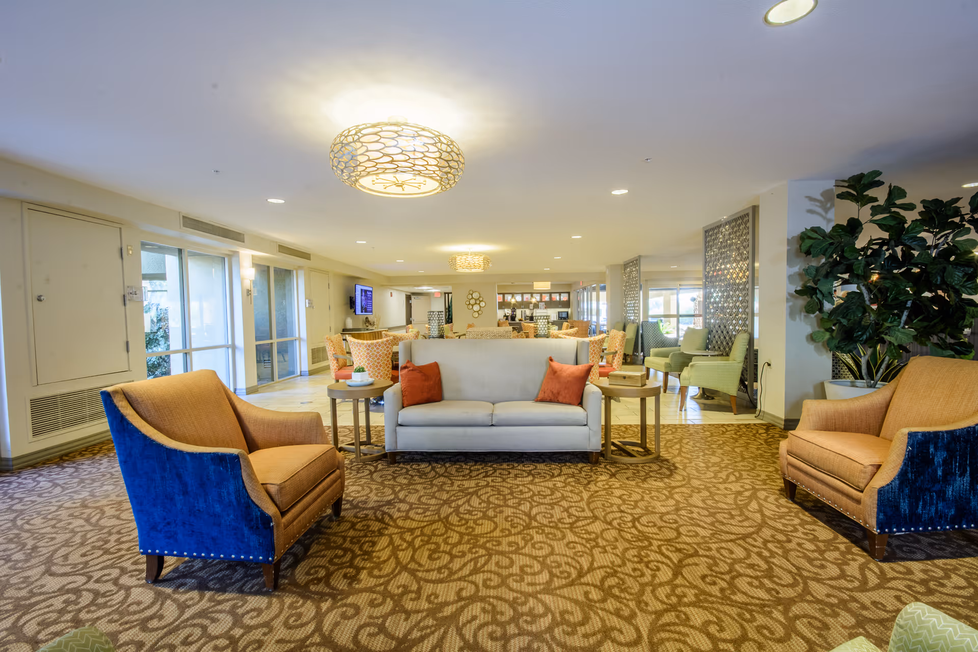 A spacious and well-lit senior living facility common area with patterned carpet flooring. The room features a beige sofa with two orange throw pillows, flanked by two armchairs with blue and tan upholstery. There are side tables with decorative items, large windows letting in natural light, and multiple seating areas with chairs and tables in the background. The ceiling has modern light fixtures, and there is a large potted plant on the right side.